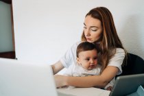 Focalisé jeune mère travaillant sur ordinateur portable tenant bébé curieux regarder vidéo drôle sur tablette tout en étant assis ensemble au bureau dans la salle de lumière — Photo de stock