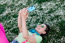 Вид сбоку радостная молодая женщина в ярком наряде лежит на пышной траве с поднятыми ногами и просматривает мобильный телефон в сельской местности — стоковое фото