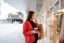 Alegre comprador femenino con bolsas de compras de papel y café para llevar de pie cerca de escaparate de la tienda en la ciudad - foto de stock
