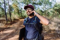 Счастливый молодой этнический мужчина турист в повседневной одежде и кепке с рюкзаком и ноутбуком разговаривает по мобильному телефону в пышной летней лесной местности — стоковое фото