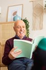 Sonriente hombre maduro sentado en silla de madera y libro de lectura mientras disfruta de fin de semana en acogedora sala de estar - foto de stock