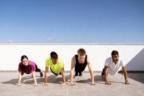 Full body group of cheerful young multiracial athletic friends in sportswear doing plank exercise on mat during workout on terrace against cloudless blue sky — Foto stock