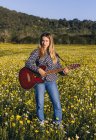 Mulher hipster jovem pensativo em pé em um prado no campo tocando guitarra durante a luz do sol de verão — Fotografia de Stock