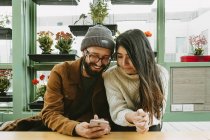 Содержание пара сидит за столом и просматривает мобильный телефон вместе во время охлаждения в кафе с разнообразными цветами горшок — стоковое фото