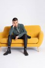 Junge hübsche männliche Modell in trendigen Klamotten sitzt auf gelben Sofa auf weißem Hintergrund im Studio — Stockfoto