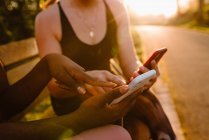 Багаторасові спортсмени в активному одязі сидять на лавці в парку і використовують мобільні телефони разом після тренування на заході сонця — стокове фото
