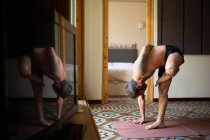 Vista laterale del maschio senza maglietta in piedi ad Ardha Baddha Padmottanasana sul tappeto mentre bilancia e pratica yoga a casa — Foto stock
