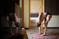 Vista lateral de un hombre sin camisa parado en Ardha Baddha Padmottanasana sobre una esterilla mientras se equilibra y practica yoga en casa - foto de stock