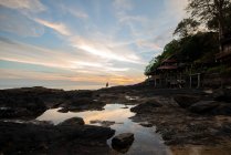 Silueta turística anónima admirando el océano sin fin contra construcciones y árboles bajo el cielo brillante al atardecer en Tailandia - foto de stock