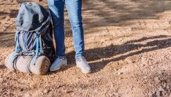 Crop escursionista anonimo in jeans e scarpe da ginnastica in piedi sulla strada sterrata vicino grande zaino nella giornata di sole — Foto stock