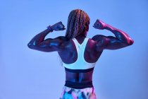 Вид сзади анонимной мускулистой афроамериканской спортсменки с потным телом, показывающей бицепсы на синем фоне — стоковое фото
