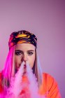 Cool hembra en traje de calle fumar cigarrillo electrónico y exhalación de humo a través de la nariz sobre fondo púrpura en el estudio con iluminación de neón rosa - foto de stock