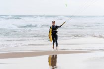 Atleta femenina en traje de neopreno con barra de control mirando a la cámara en la orilla arenosa contra el océano espumoso después de practicar kitesurf - foto de stock