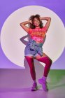 Full body fit African American female dancer in shorts dancing with arms raised and sticking tongue out looking away while standing in neon lights in studio — Stock Photo