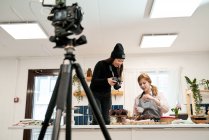 Женщина фотографирует шоколадные кексы на цифровой камере против блогера, разговаривающего во время приготовления пищи на кухне — стоковое фото