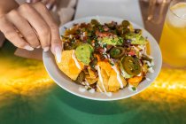 Top view of crop unrecognizable female eating delicious traditional Mexican nachos with jalapeno chili pepper topped with cheese and guacamole sauce sitting at table in restaurant — Stock Photo