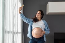 Sonriendo hembra embarazada tocando el vientre mientras está de pie en la habitación en casa y tomando selfie con teléfono móvil - foto de stock