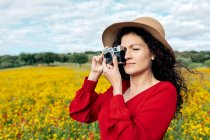 Улыбающаяся женщина в шляпе фотографирует на винтажную камеру на лугу под облачным небом — стоковое фото