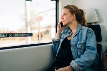Jovem mulher casual em jaqueta de ganga ter telefonema olhando para longe na janela do trem enquanto viaja — Fotografia de Stock