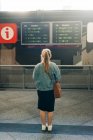 Vue arrière de la femelle veste en denim et avec sac regardant l'horaire des trains à bord debout sur la gare — Photo de stock