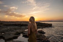 Вид сбоку на молодую женщину, стоящую на берегу на фоне синего волнистого моря на закате и отводящую взгляд — стоковое фото