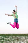 Ganzkörperentzückte junge Frau in kurzen Hosen und leuchtend rosa Strumpfhosen hüpft fröhlich mit erhobenem Arm auf einer grünen Lichtung in der Natur — Stockfoto