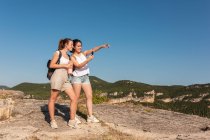 Happy young female travelers in summer clothes using compass together while standing on lush sunny hilly terrain — Stock Photo