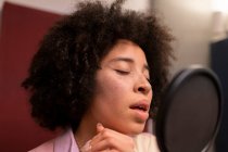 Чорна співачка виконує пісню проти мікрофона з поп-фільтром, стоячи і закриваючи очі на звуковій студії — стокове фото