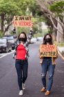 Этнические женщины в масках держат плакаты с протестами против расизма на городской улице и смотрят в камеру — стоковое фото