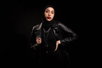 Attraente giovane donna islamica vestita di nero con giacca di pelle e hijab che guarda delicatamente la fotocamera sullo studio nero — Foto stock