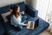 Положительная беременная женщина сидит на мягком диване с ноутбуком и пьет горячий напиток — стоковое фото
