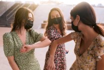 Зміст найкращих друзів-жінки в декоративних сукнях і масках для обличчя, які торкаються ліктів, дивлячись один на одного в місті під час пандемії коронавірусу — стокове фото