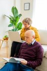 Mann mittleren Alters liest Buch und reife Frau surft Laptop, während sie es sich im gemütlichen Wohnzimmer zu Hause gemütlich macht — Stockfoto