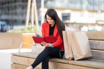 Smiling cliente feminino sentado no banco com sacos de papel e livro de leitura após compras bem sucedidas na cidade — Fotografia de Stock