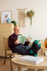 Lächelnder älterer Mann sitzt auf Holzstuhl und liest Buch, während er das Wochenende im gemütlichen Wohnzimmer genießt — Stockfoto
