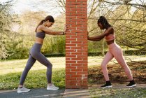 Side view of young multiethnic sportswomen in activewear working out between rough post while looking down in city park — Stock Photo