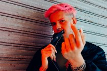 Jeune homme homosexuel avec tatouage et cheveux roses dans des vêtements de dessus élégants regardant la caméra contre les murs altérés — Photo de stock