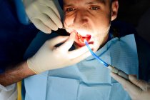 Crop anonymous orthodontist in latex gloves with coworker checking up teeth of mature man looking forward in hospital — Stock Photo