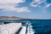 Empty white chairs on deck of cruise boat sailing in blue sea water with mountain on shore — Stock Photo