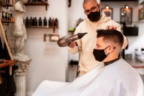 Estilista en máscara textil con secador de pelo contra hombre en capa en sillón en barbería - foto de stock