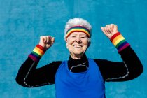 Захоплена старша жінка в спортивному одязі, що святкує перемогу з згорнутими кулаками та піднятими руками на синьому фоні в студії — стокове фото