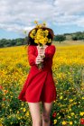 Анонімні жінки в капелюсі покривають обличчя квітучими жовтими квітами в сільській місцевості під хмарним небом — стокове фото