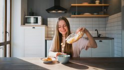 Fröhliche Frau gießt knuspriges Müsli in Schüssel am Tisch mit leckeren Haferkeksen mit Schokochips zum Frühstück zu Hause — Stockfoto
