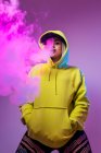 Von unten selbstbewusste Hipsterin im Kapuzenpulli, E-Zigarette rauchend im Studio auf rosa Hintergrund und in die Kamera blickend — Stockfoto