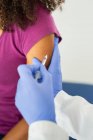 Especialista médica feminina irreconhecível em uniformes de proteção e luvas de látex vacinando uma paciente afro-americana anônima na clínica durante o surto de coronavírus — Fotografia de Stock