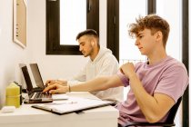 Вид збоку концентрованих молодих багатоетнічних колег-чоловіків у повсякденному вбранні, які працюють дистанційно на ноутбуках, сидячи за столом вдома — стокове фото