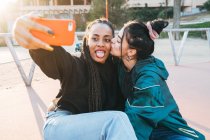 Jeune femme homosexuelle embrasser noir bien-aimé avec la langue tout en prenant autoportrait sur téléphone portable en ville dans le dos éclairé — Photo de stock