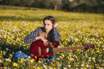 Thoughtful young hipster woman sitting on a meadow in the countryside writing songs on notebook and playing guitar during summer sunlight — Stock Photo