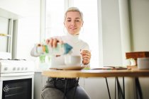 Astrólogo feminino sorridente derramando leite de caixa em bebida quente à mesa em casa no dia ensolarado — Fotografia de Stock