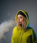 Confident female hipster in hoodie smoking e cigarette in studio on gray background and looking away — Stock Photo